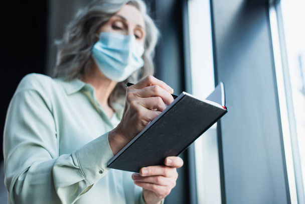 Low angle view van wazig zakenvrouw in medisch masker schrijven op notebook in het kantoor  - Foto, afbeelding