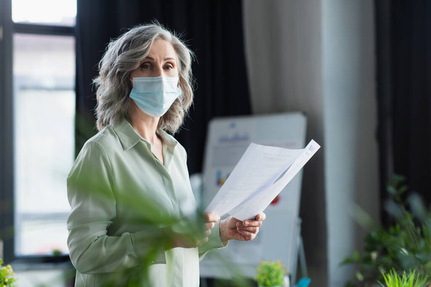 Mujer de negocios de pelo gris con máscara médica sosteniendo documentos y mirando a la cámara en la oficina  - Foto, Imagen