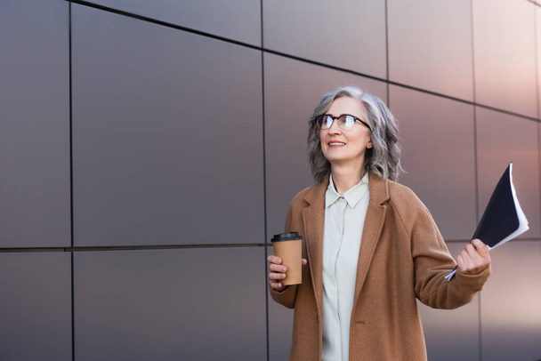 Ευτυχισμένη επιχειρηματίας σε παλτό κρατώντας καφέ για να πάει και το φάκελο χαρτί κοντά κτίριο σε εξωτερικούς χώρους  - Φωτογραφία, εικόνα