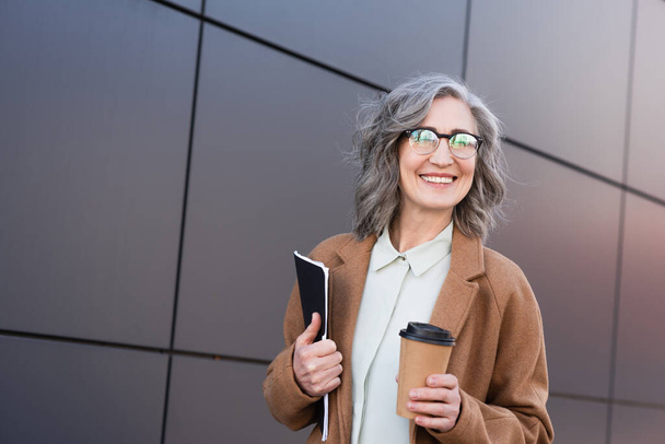 Middle aged businesswoman in coat and eyeglasses holding paper folder and coffee to go outdoors  - Foto, immagini