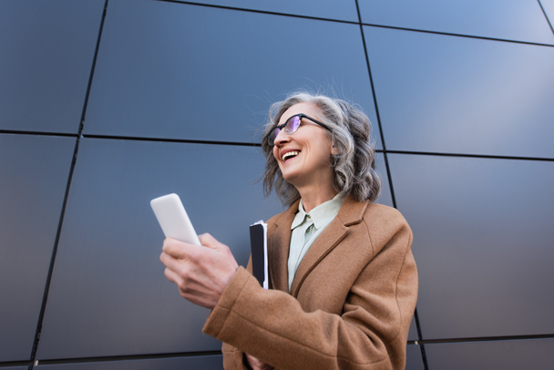 Visão de baixo ângulo de empresária alegre no casaco segurando celular e pasta de papel perto do edifício ao ar livre  - Foto, Imagem