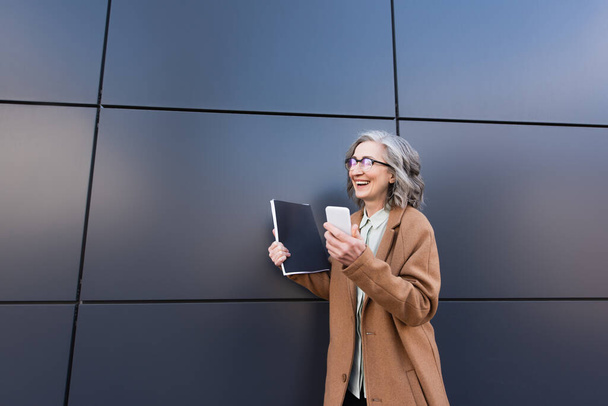 Positive businesswoman in coat holding smartphone and paper folder near building outdoors  - Photo, Image