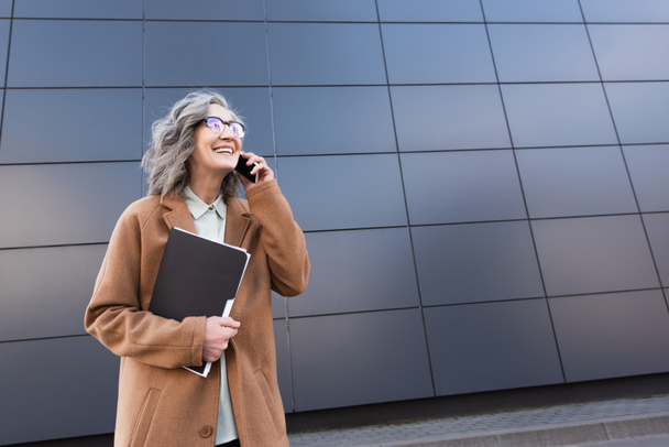 Cheerful grey haired businesswoman with papers talking on cellphone on urban street  - Foto, afbeelding