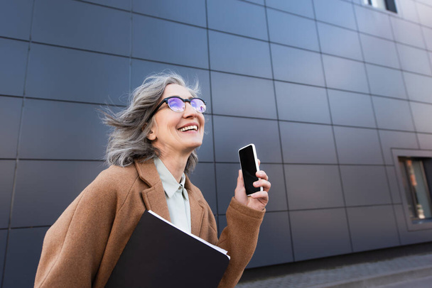 Szczęśliwa bizneswoman w płaszczu trzymając teczkę papieru i telefon komórkowy z pustym ekranem na ulicy miejskiej  - Zdjęcie, obraz
