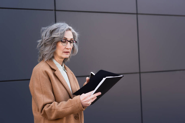 Madura mujer de negocios en gafas y abrigo sosteniendo la carpeta de papel y el teléfono celular al aire libre  - Foto, imagen