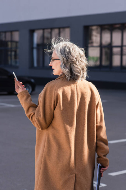 Side view of mature businesswoman in coat holding documents and using cellphone on urban street  - Fotografie, Obrázek