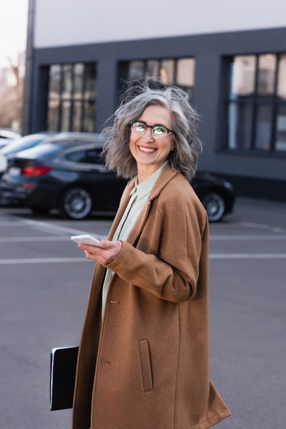 Mature businesswoman in coat holding mobile phone and paper folder on urban street  - Fotografie, Obrázek