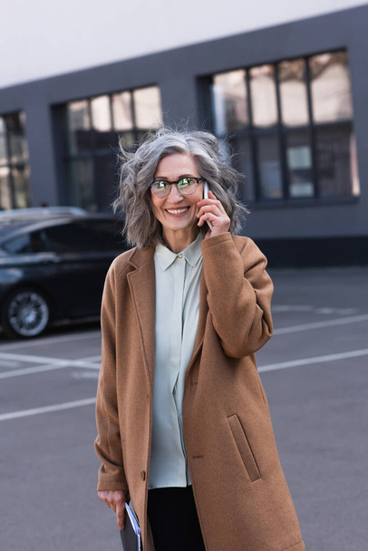 Femme d'affaires mature gaie en manteau parlant sur son téléphone portable et tenant des documents dans la rue urbaine  - Photo, image