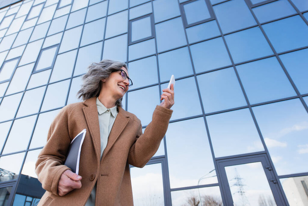 Low angle view of cheerful businesswoman in coat holding paper folder and looking at cellphone on urban street  - Photo, image