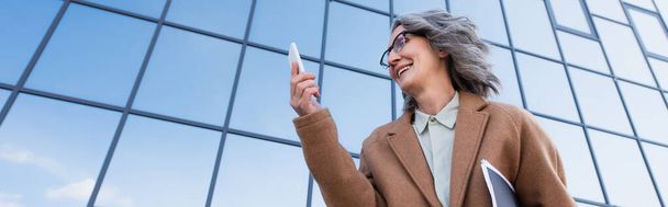 Low angle view of businesswoman in coat holding papers while looking at cellphone outdoors, banner  - Fotografie, Obrázek