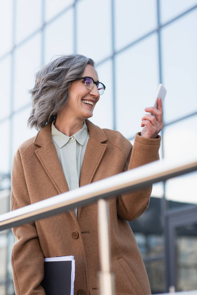 Щаслива бізнес-леді в пальто, що тримає документи і мобільний телефон біля розмитих перил на відкритому повітрі
  - Фото, зображення