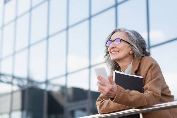 Mature businesswoman in eyeglasses and coat holding cellphone and paper folder near railing outdoors  - Фото, изображение