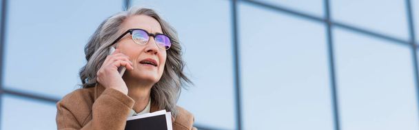 Empresária de cabelos grisalhos em óculos falando no celular e segurando pasta de papel ao ar livre, banner  - Foto, Imagem