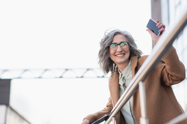 Rijpere zakenvrouw in jas houden smartphone met blanco scherm en papieren map in de buurt railing buiten  - Foto, afbeelding