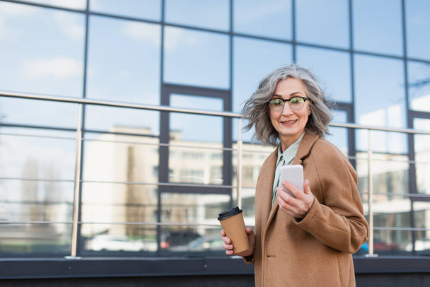 Usmívající se podnikatelka v kabátě pomocí smartphonu a držení kávy jít na městské ulici  - Fotografie, Obrázek
