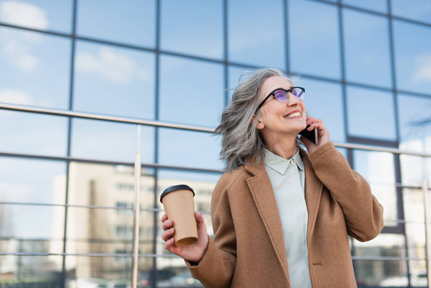 Happy senior businesswoman talking on smartphone and holding paper cup on urban street  - Photo, Image