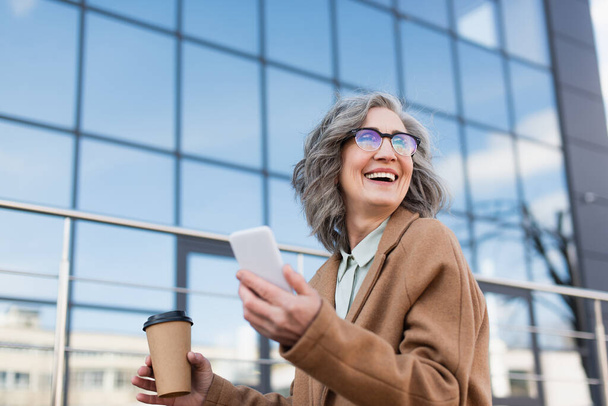 Joyful businesswoman in coat holding blurred smartphone and paper cup outdoors  - Fotó, kép