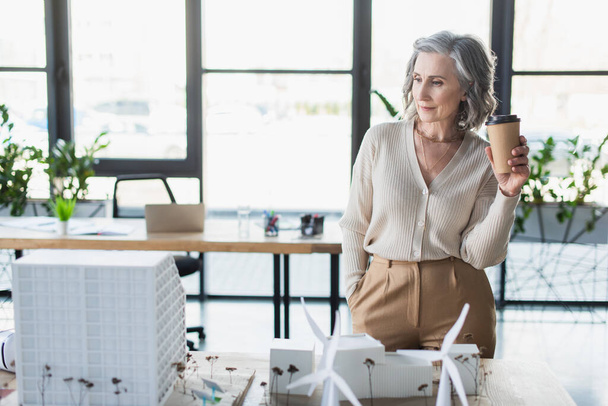 Reife Geschäftsfrau hält Kaffee in der Nähe von Modellen von Bürogebäuden  - Foto, Bild