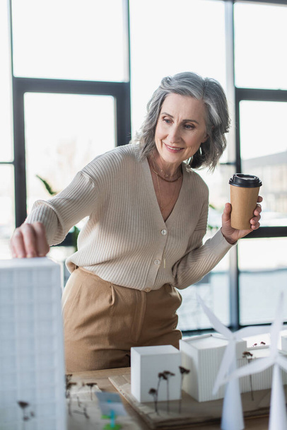 Mujer de negocios de pelo gris sonriente sosteniendo una taza de papel cerca de modelos borrosos de edificios en la oficina  - Foto, Imagen