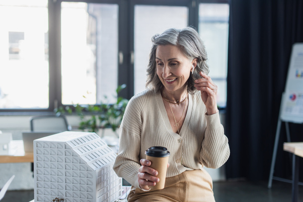 Lächelnde Geschäftsfrau mit Kopfhörer hält Pappbecher in der Nähe von Modell des Bürogebäudes  - Foto, Bild