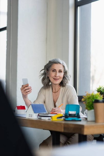 Businesswoman smiling at camera while holding smartphone near models of houses in office  - Фото, изображение