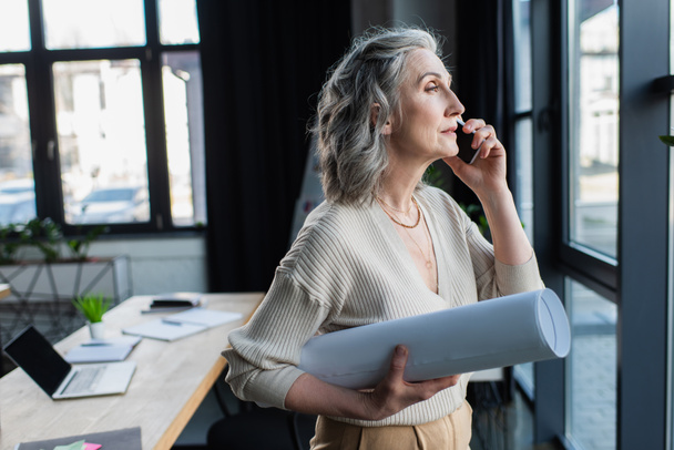 Mature businesswoman with blueprint talking on smartphone in office  - Photo, Image