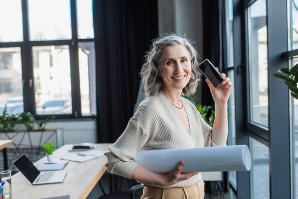 Cheerful mature businesswoman holding smartphone with blank screen and blueprint in office  - Fotó, kép