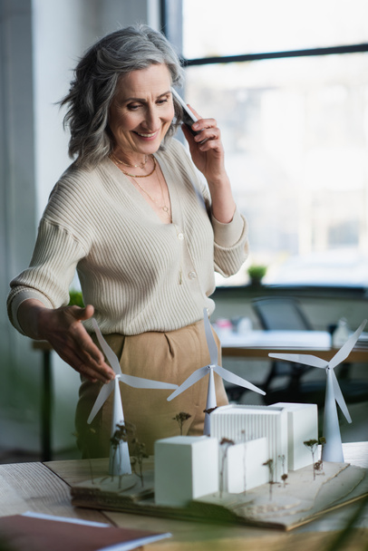 Dojrzała bizneswoman rozmawia na smartfonie w pobliżu modeli budynków i folderu papierowego w biurze  - Zdjęcie, obraz