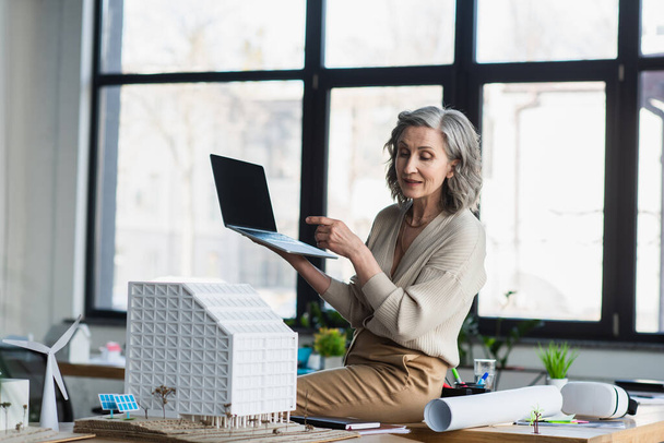 Femme d'affaires ayant appel vidéo sur ordinateur portable près du plan directeur et modèle de bâtiment dans le bureau  - Photo, image