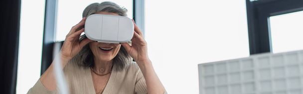 Šedovlasá podnikatelka používající sluchátka virtuální reality v blízkosti rozmazaného modelu budovy v kanceláři, banner  - Fotografie, Obrázek