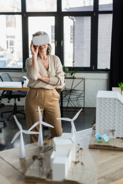 Χαρούμενη επιχειρηματίας σε εικονική πραγματικότητα ακουστικά στέκεται κοντά στο έργο με τα μοντέλα των κτιρίων στο γραφείο  - Φωτογραφία, εικόνα