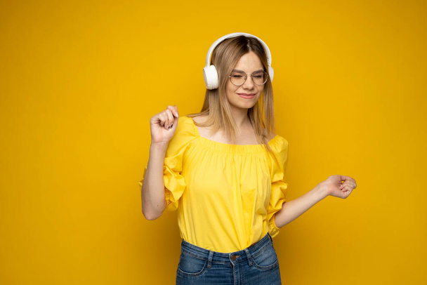 Happy girl in a glasses and yellow shirt dancing and listening to the music isolated on a yellow background. - Fotó, kép