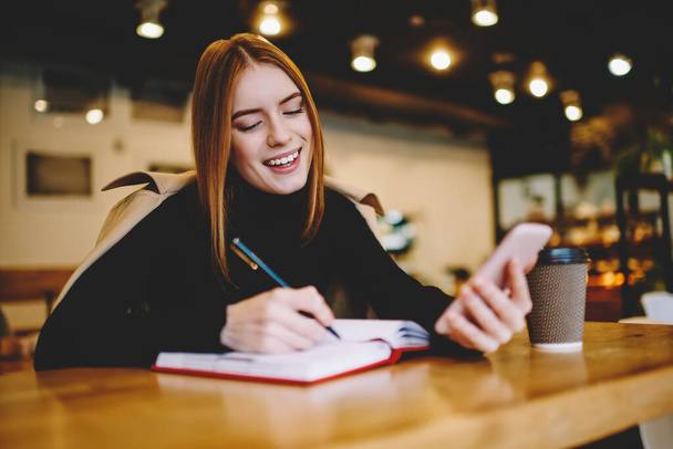 Smiling female in casual clothes writing thoughts in notebook and using smartphone while chilling in cafe with cozy interior in daytime - Valokuva, kuva
