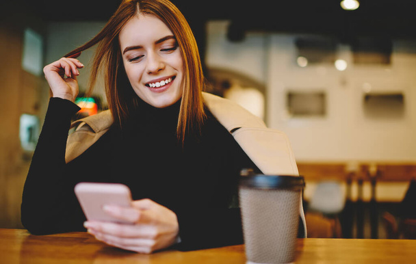 Smiling female in casual clothes touching hair and using smartphone while sitting at table with cup of coffee in cozy cafe - Valokuva, kuva