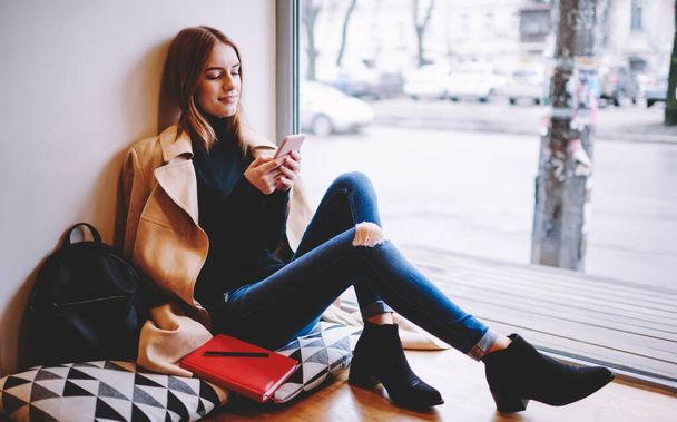 Side view of charming female in stylish clothes sitting on windowsill near large window and using smartphone while chilling in cafe in daytime - Foto, Imagem