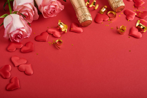 Pink roses flowers, champagne, gift, golden ribbons and confetti red hearts on red background. Top view flat lay with space for your greetings. Valentines day background and greeting card. - Photo, Image