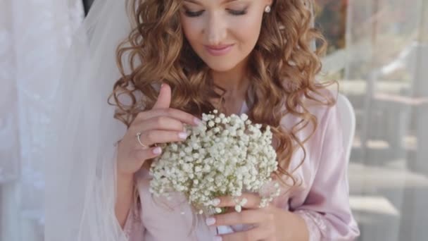 Een tedere jonge bruid in een sluier houdt een boeket zigeunerbloemen in haar handen en raakt de witte bloemen met haar hand aan. Langzame beweging - Video