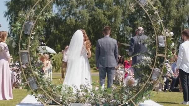 Widok z tyłu panny młodej i pana młodego stojących przy łuku kwiatów przed gośćmi i słuchających przemówienia podczas ceremonii ślubnej - Materiał filmowy, wideo