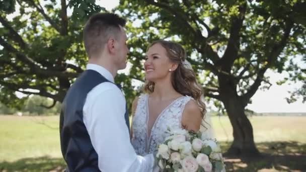 幸せな新婚夫婦は木の下に立ち、新郎は美しいレースのドレスに花束を手に花嫁の鼻にキスをします。スローモーション - 映像、動画