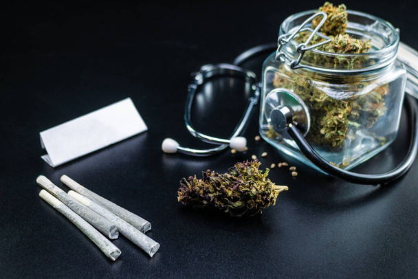 Сухая медицинская марихуана в банке со стетоскопом на черном фоне - Фото, изображение