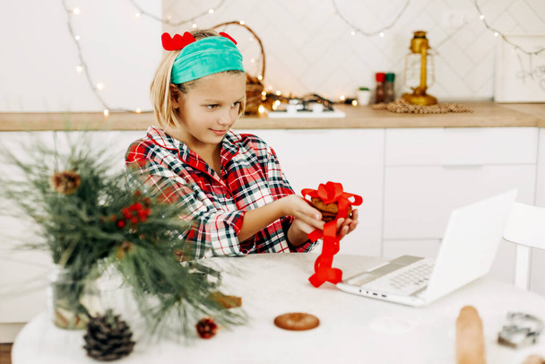 Ein Mädchen, das zu Weihnachten zu Hause in der Küche sitzt, gratuliert seinen Freunden oder Großeltern per Videoübertragung, zeigt ihnen ein Geschenk aus selbst gebackenen Plätzchen - Foto, Bild