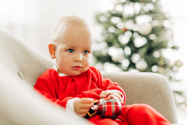 Ребенок в костюме Санта Клауса сидит в кресле рядом с елкой в канун Рождества - Фото, изображение