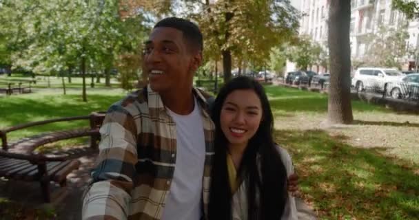 Happy multiracial young loving couple actively walking in the park and recording video or having video call on mobile phone in the outstretched of a handsome African guy hugging his pretty girlfriend - Footage, Video