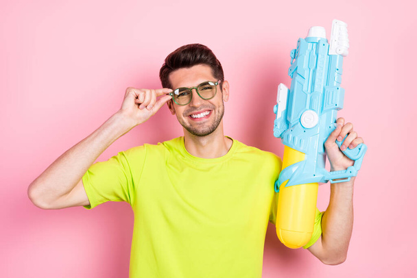 Photo portrait man wearing bright t-shirt glasses keeping water gun smiling isolated pastel pink color background - Foto, Imagem