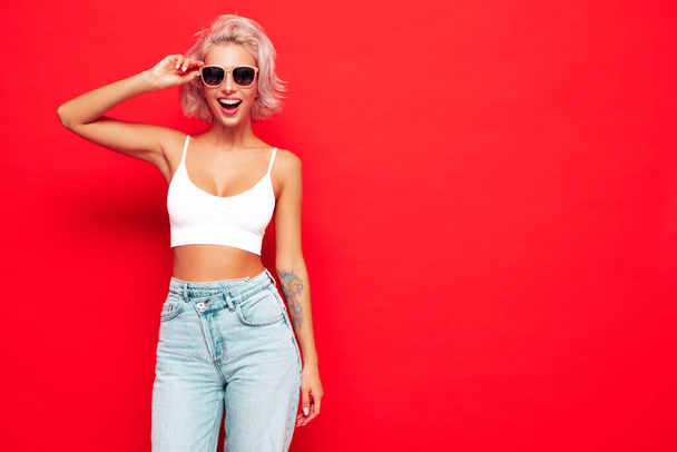 Young beautiful smiling female in trendy summer clothes. Sexy carefree woman posing near red wall in studio. Positive blond model having fun and going crazy. Cheerful and happy. In sunglasses - Fotoğraf, Görsel