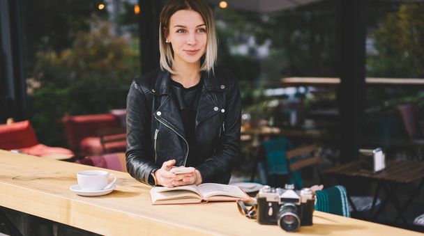 Young woman sitting in modern cafe at table with cup of drink and photo camera and with open book while checking notification on smartphone while looking away - Photo, Image