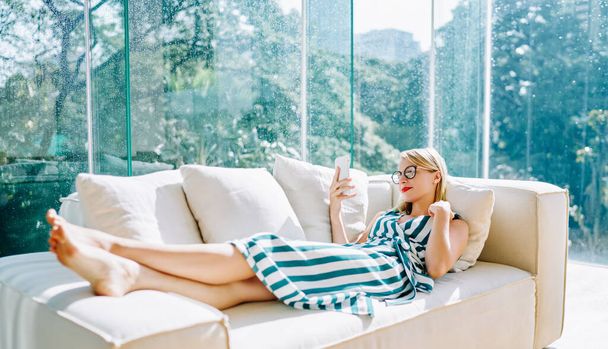 Side view of young female in smart casual and eyeglasses lying on white sofa with cushions and reading book on smartphone in glass terrace - Foto, Imagem