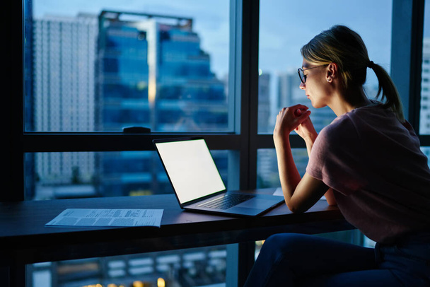 Zijaanzicht van gerichte vrouw met gevouwen handen zitten aan tafel met geopende laptop tijdens het werken aan business project in de avond - Foto, afbeelding