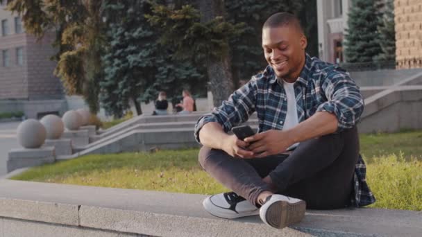 Feliz chico afroamericano amigable joven sentado en la ciudad en la calle al aire libre mirando el teléfono móvil mira a la distancia saludando saludo amigo invitando a venir aquí bienvenida invitan gesto - Metraje, vídeo