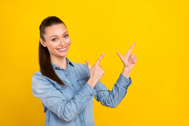 Photo of funny cute young woman wear denim shirt smiling pointing two fingers empty space isolated yellow color background - Foto, imagen
